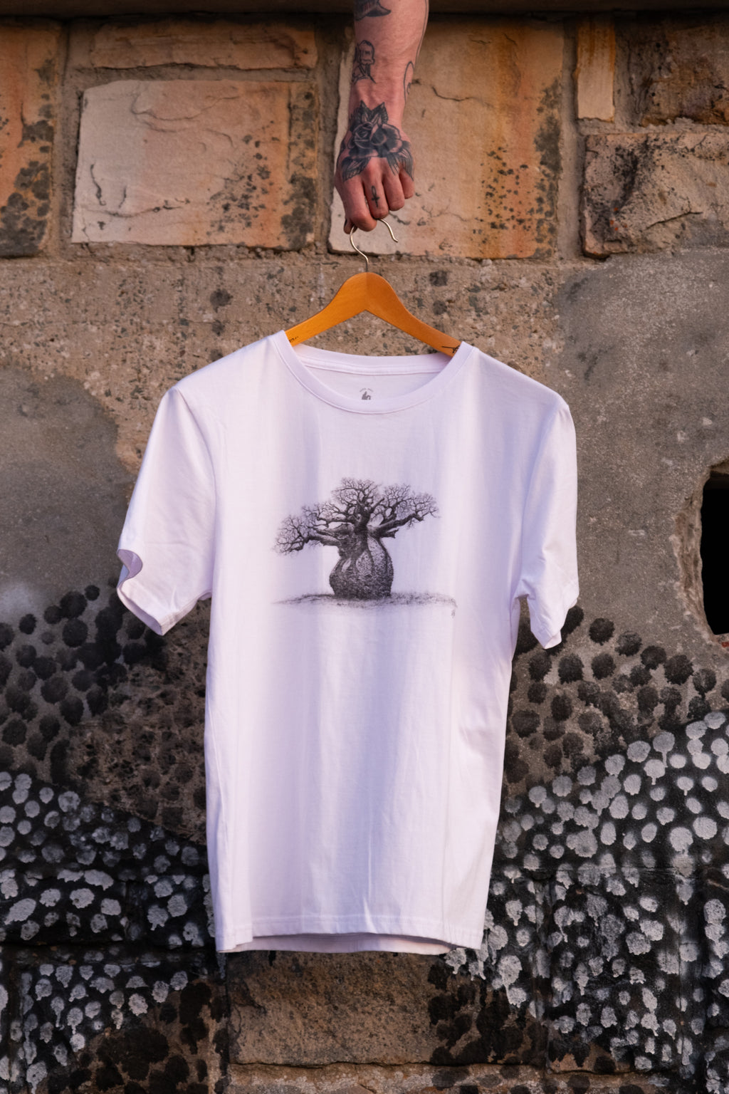 White t-shirt with a baobab tree design on a hanger against a textured stone wall.