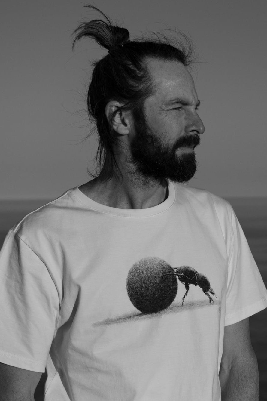 Man wearing a white t-shirt with a graphic design of a dung beetle and ball on a beach.